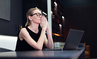Stylish elegant woman  in glasses praying. Laptop on the table in modern office. Bussiness concept.