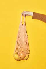 Vibrant close up of hand holding eco shopping bag with oranges against yellow background, conscious consumption and vegan concept