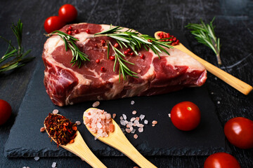 Close-up piece of beef meat ribeye with spoons of Himalayan salt and grilling spices, tomato and rosemary. Side view. BBQ ingredients.