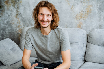 Fototapeta na wymiar A guy in a gray T-shirt sits on the couch at home with a big smile on his face. A cheerful young man, long hair, a hipster beard mustache.