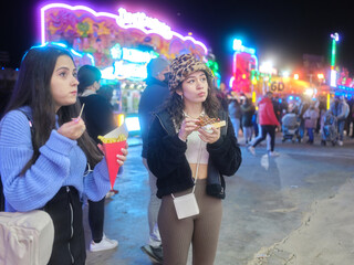 Distracted women eating french fries and waffle standing on a night fair