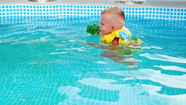 The child swims in the pool in a yellow vest. A little boy swims in a blue pool and plays with a toy. Summertime