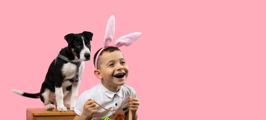 Picture of a smiling boy wearing bunny ears holding basket with painted eggs and his lovely dog sitting behind him on a chair, together posing on pink background and copy space.