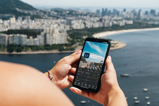 Girl holding smartphone with Adobe Photoshop Lightroom app on screen. City and bay with some boats in the background. Rio de Janeiro, RJ, Brazil. March 2022