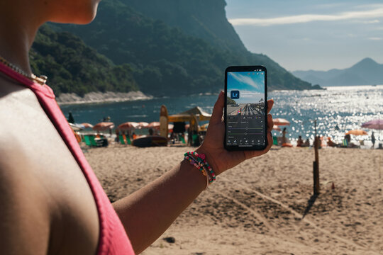 Girl on the beach holding a smartphone with Adobe Photoshop Lightroom app on the screen. Rio de Janeiro, RJ, Brazil. March 2022