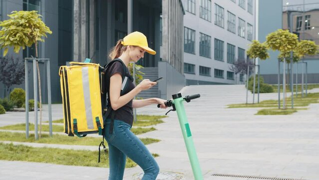Female deliveryman of takeaway with backpack isothermal case box. Close-up of courier looks at delivery address on phone. Smiling woman with thermal backpack looking at smartphone