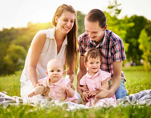 Happy family with young, millennial parents and two baby girls sitting on a picnic blanket and  having fun together - Outdoor lifestyle family concept