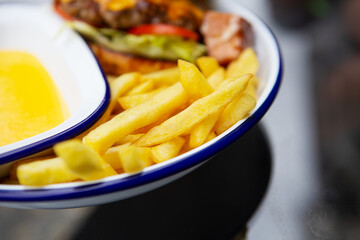 The concept of fast food. French fries lie on a meta plate along with cheddar cheese sauce and a hamburger in the background.