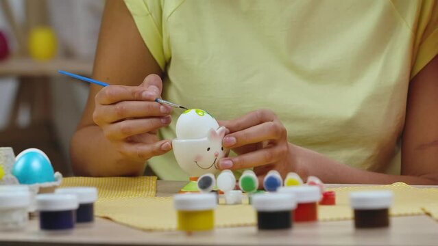 Mom teaches her daughter to paint eggs with paints and brush. African American woman and little girl with bunny ears are sitting at table in decorated room. Happy easter. Slow motion. Close up.