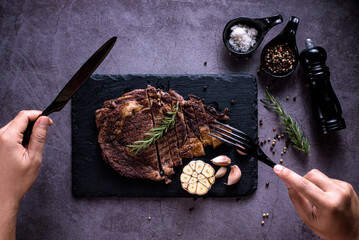 Sliced beef ribeye steak, grilled with 4seasons pepper garlic-rosemary, on stone plate dark and grey background, hands holding a knife and fork, top view
