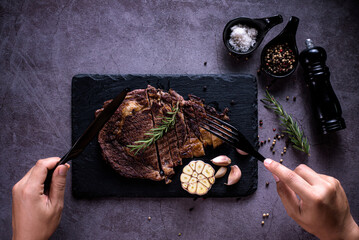 Sliced beef ribeye steak, grilled with 4seasons pepper garlic-rosemary, on stone plate dark and grey background, hands holding a knife and fork, top view