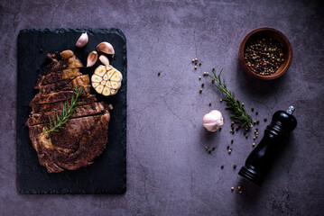 Sliced beef ribeye steak, grilled with 4seasons pepper garlic-rosemary, on stone plate dark and dark grey background, knife and fork black, top view