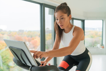 Home cycle workout Asian fit athlete woman training on stationary bike doing online cycling exercise class biking during livestream on screen.