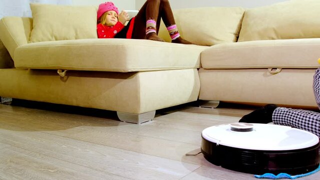 Little girl plays with mobile phone and sister looks at robot vacuum cleaner on floor in living room. Technology for effective housekeeping