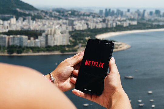 Girl holding smartphone with Netflix app on screen. City and bay with some boats in the background. Rio de Janeiro, RJ, Brazil. March 2022