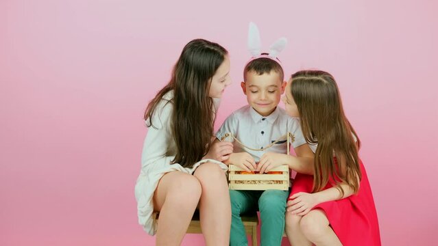 Three children sitting together on a chair, boy wearing rabbit eras on head holding basket with painted eggs and sister kissing him on cheeks.