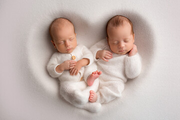 Tiny newborn twin boys in white bodysuits on a white background. Newborn twins sleep next to their brother on the background of the heart. Two newborn twin boys hugging each other. - obrazy, fototapety, plakaty