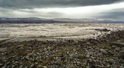 A wind-swept lake rolling in on a rocky shoreline with a stormy sky and mountains in the background.