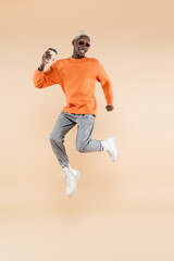 full length of happy african american man in sunglasses levitating while holding paper cup on beige