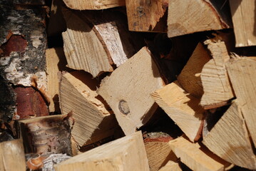 A neatly folded pile of firewood. The sun partially illuminates a neatly stacked stack of firewood. Birch chocks are stacked perpendicular to each other forming a wall.