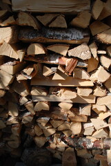 A neatly folded pile of firewood. The sun partially illuminates a neatly stacked stack of firewood. Birch chocks are stacked perpendicular to each other forming a wall.