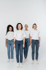 Full length of smiling multiethnic women in white t-shirts standing on grey background.