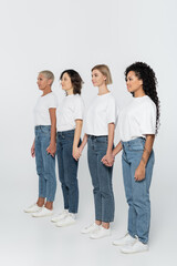 Fototapeta premium Multiethnic women holding hands and looking away on grey background.