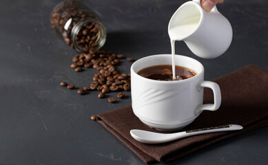 Milk is poured into a cup of coffee on a gray table, in the background a jar with coffee beans
