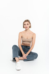 Blonde woman in top and jeans sitting on grey background.