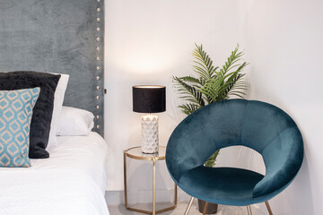 Bed with blue upholstered headboard with blue and white cushions, blue velvet upholstered chair and lamp on the bedside table