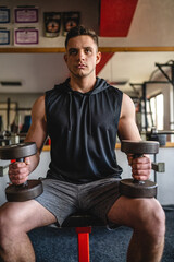 One man young adult caucasian male bodybuilder training arms bicep flexing muscles with dumbbell while sitting in the gym wearing shirt real people copy space front view waist up