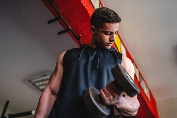 One man young adult caucasian male bodybuilder training arms bicep flexing muscles with dumbbell while standing in the gym wearing shirt real people copy space front view waist up