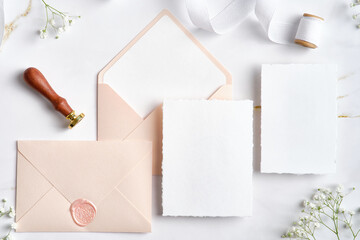 Blank white invitation cards with pink envelopes, gypsophila branches, wax seal stamp, silk ribbon. Wedding stationery set. Flat lay, top view, copy space.