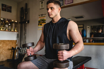 One man young adult caucasian male bodybuilder training arms bicep flexing muscles with dumbbell while sitting in the gym wearing shirt real people copy space front view waist up