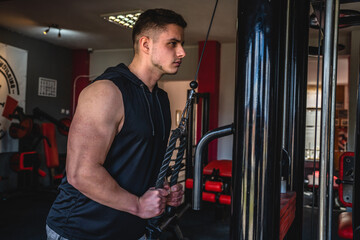 Naklejka premium One man side view caucasian male bodybuilder at gym wearing black shirt workout using rope and cable weights for arms exercise triceps training copy space waist up dark photo real people amateur