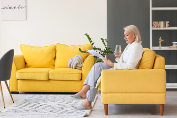 Mature woman with glass of water reading newspaper in armchair at home