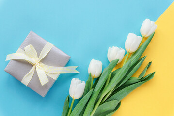 Gift box and white tulips on two toned blue and yellow background. Spring holidays concept. Top view, flat lay, copy space