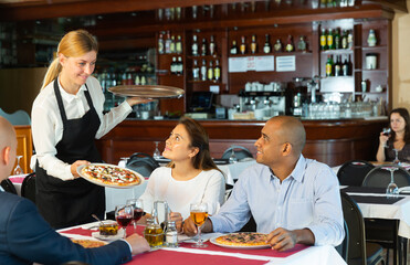 Polite smiling female waiter bringing ordered pizza to friends