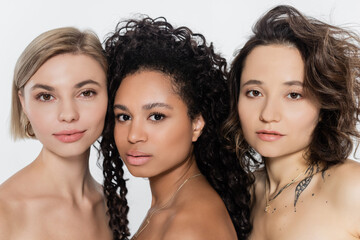 Young multiethnic women with naked shoulders looking at camera isolated on grey.