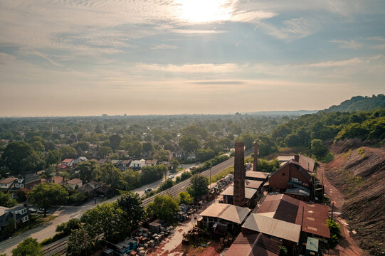 An aerial view of a hazy sunny morning over a brickyard and residential neighbourhood filled with lush green trees under a blue sky during summer, in Hamilton, Ontario, Canada.