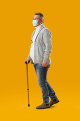 Senior man in medical mask with walking stick on yellow background