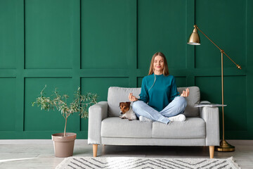 Young woman with Jack Russel terrier meditating on grey sofa near green wall