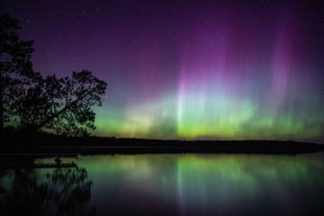 Northern lights erupt above a lake in Northern Minnesota in the dark sky overhead shining a rainbow...