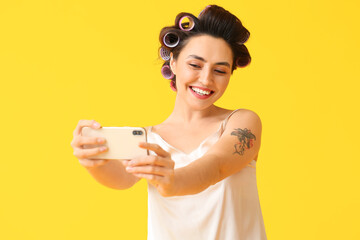 Beautiful young woman in hair curlers taking selfie on yellow background