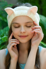 Beautiful little teenage girl with funny cat headband is smiling front of tropical leaves background. Portrait of cute little child enjoying skin care procedures