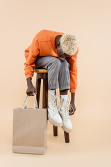 full length of blonde african american man in orange sweatshirt holding shopping bag and sitting on chair on beige