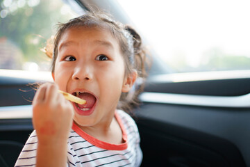 Little Asian young girl staying in the car and eating fried potato chips.