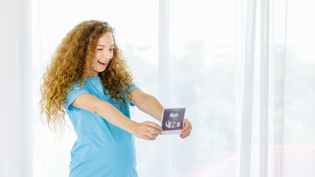 Caucasian young beautiful long curly hairstyle healthy pregnant mother in casual outfit standing holding showing baby ultrasound Xray film picture sonogram in hands in front of curtain background