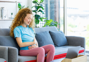 Caucasian happy pretty long curly hair healthy pregnancy mother in casual outfit sitting smiling on cozy sofa touching rubbing tummy belly watching online streaming via laptop computer in living room