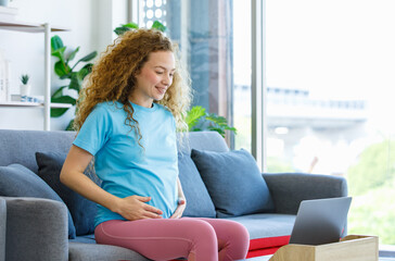 Obraz premium Caucasian happy pretty long curly hair healthy pregnancy mother in casual outfit sitting smiling on cozy sofa touching rubbing tummy belly watching online streaming via laptop computer in living room